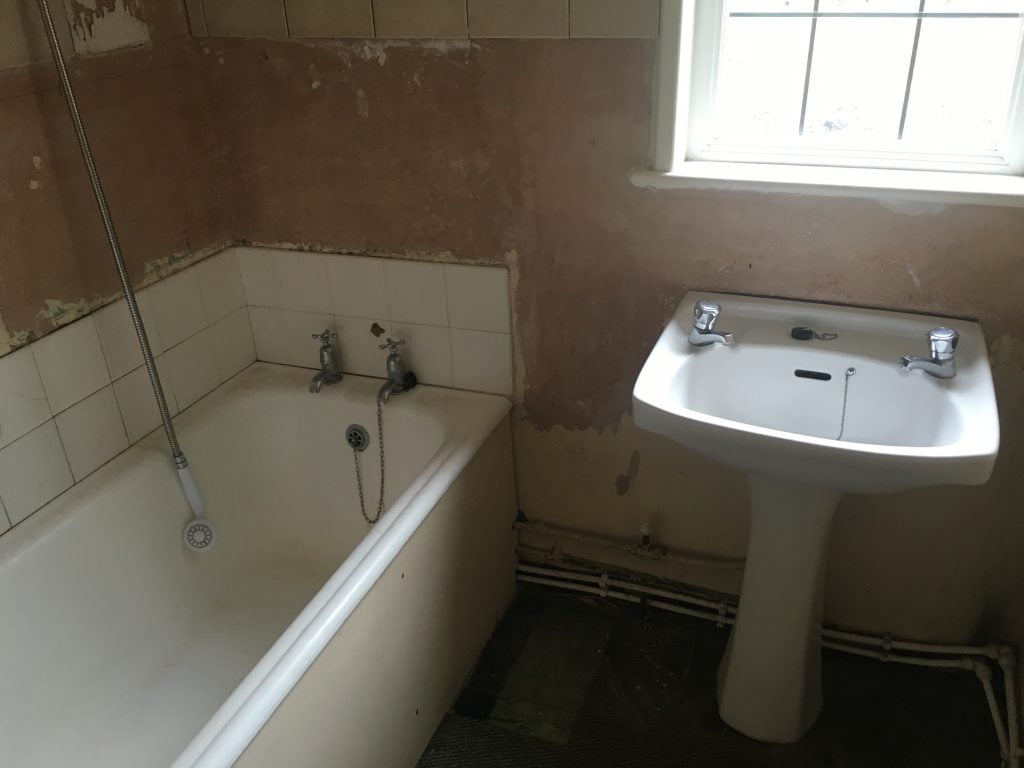 White bath and sink with shower head hanging down surrounded by bare plastered walls