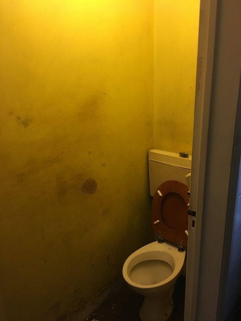 Old, dirty toilet cubicle with white basin and brown wooden seat